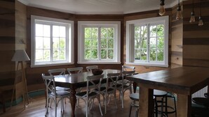 Ein Essbereich mit Holztisch und Stühlen, großen Fenstern mit Blick ins Grüne und einer Decke mit freiliegenden Balken und Pendelleuchten.