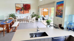 Een moderne keuken met een centraal eiland, een eethoek met een tafel en stoelen, en een zithoek met een bank en een groot raam.