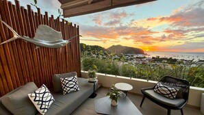 Een balkon met uitzicht op de ondergaande zon boven de oceaan, een hangmat, een bank met kussens, een kleine tafel en een stoel.