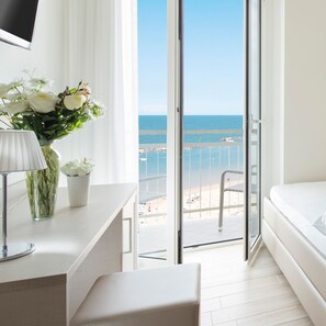 Habitación de hotel con balcón y vistas al mar, una cama, un escritorio con lámpara y un florero con flores.