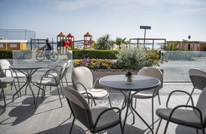 Un área con mesas y sillas redondas al aire libre, un parque infantil al fondo y vista al mar.