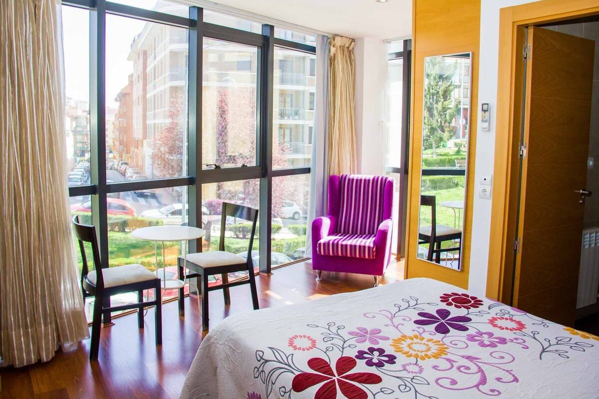 A hotel room with large windows, a bed with a floral bedspread, a purple chair, and a small table.