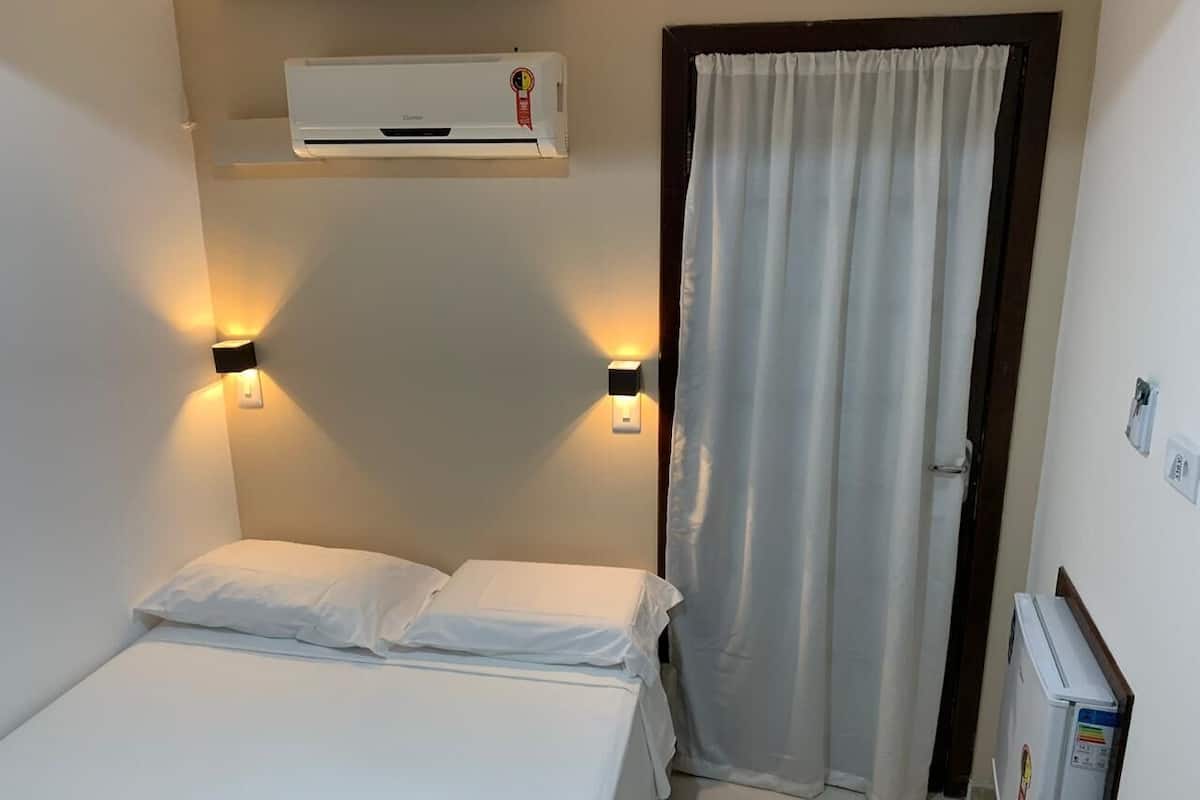 A small, clean room with a bed, two wall-mounted lights, an air conditioner, a curtain, and a small refrigerator.