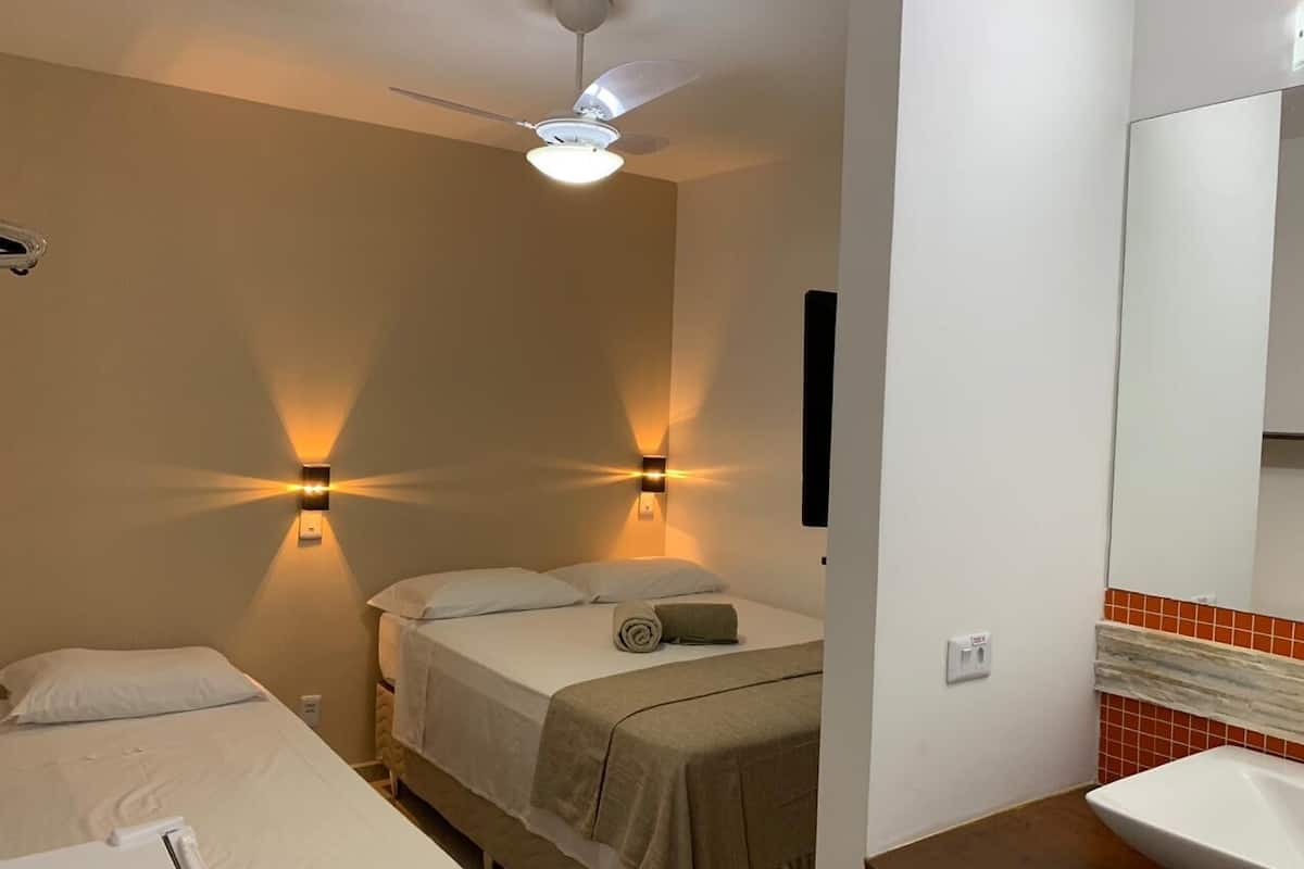 A hotel room with two beds, a ceiling fan, wall-mounted lights, a bathroom with a sink, and a mirror.