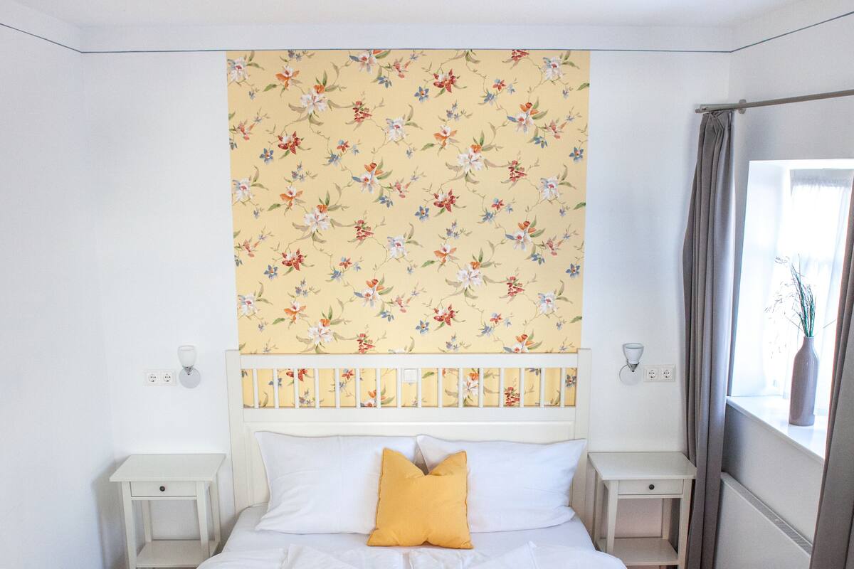 A bedroom with a bed, a nightstand, a window with curtains, and a floral-patterned wall.