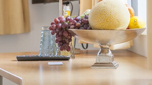 A table with a fruit bowl containing grapes, a melon, and a lemon, a remote control, and a glass of water. - Hotel am Markt (Hungen)