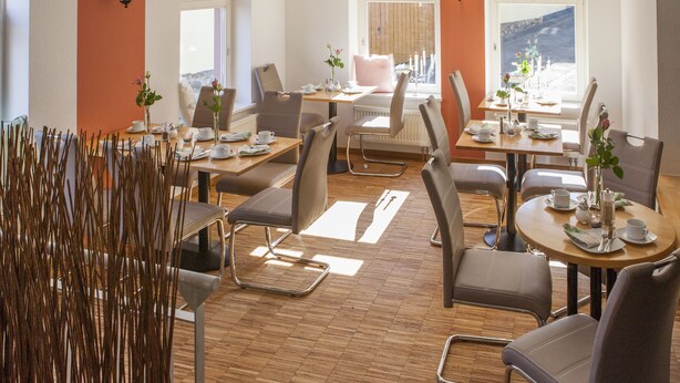 A dining area with multiple tables and chairs, a view of the outdoors through large windows, and a decorative plant arrangement. - Hotel am Markt (Hungen)