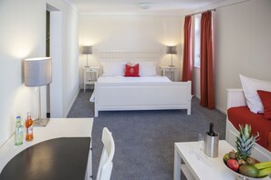 Una habitación de hotel con una cama, mesitas de noche, un escritorio, una silla, una pequeña mesa con fruta y una botella, y una lámpara.