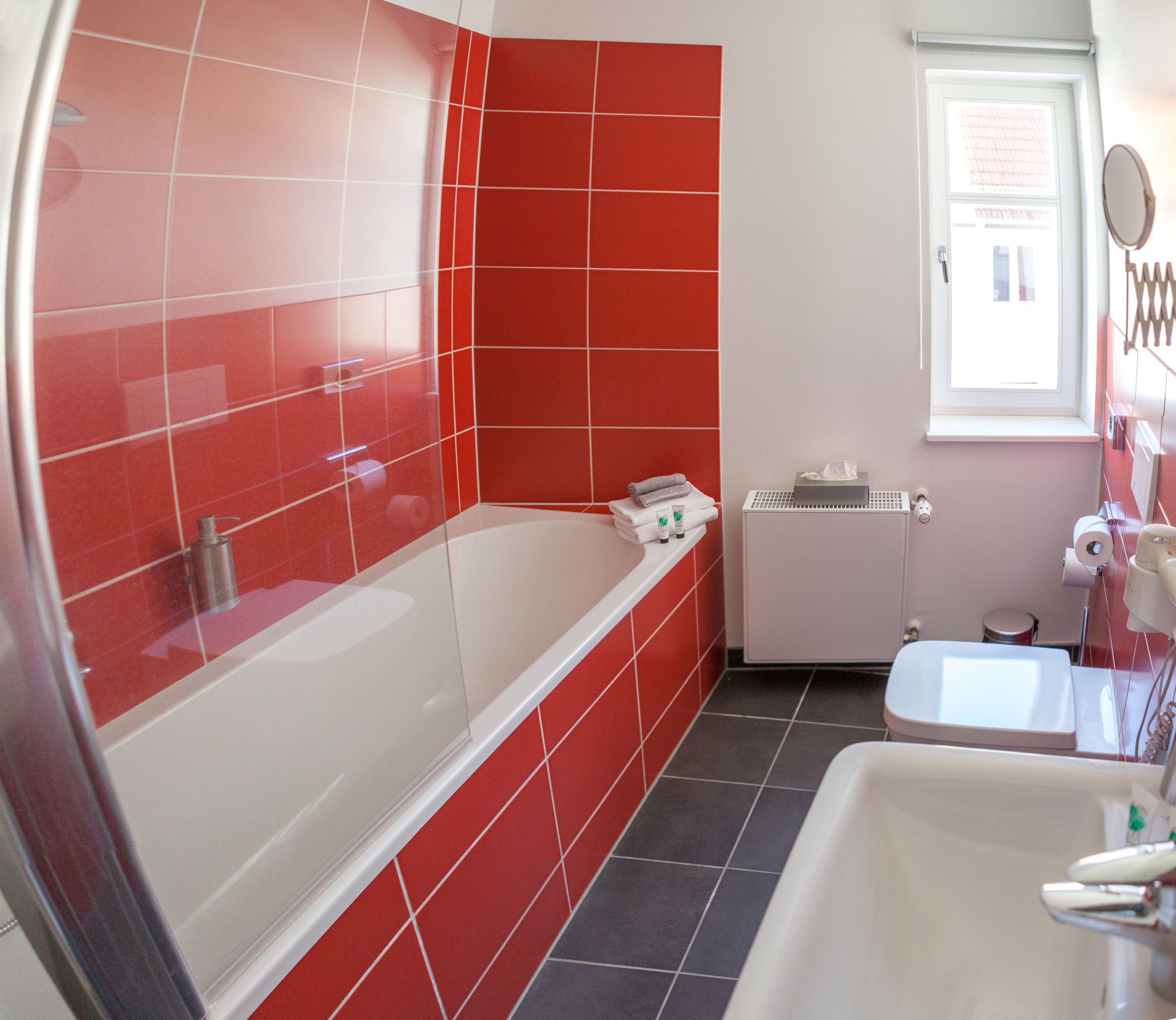Un baño con azulejos rojos, una bañera blanca, un lavamanos blanco y una ventana con espejo.