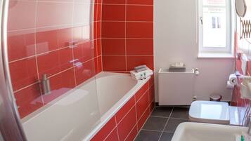 Un baño con azulejos rojos, una bañera blanca, un lavamanos blanco y una ventana con espejo.
