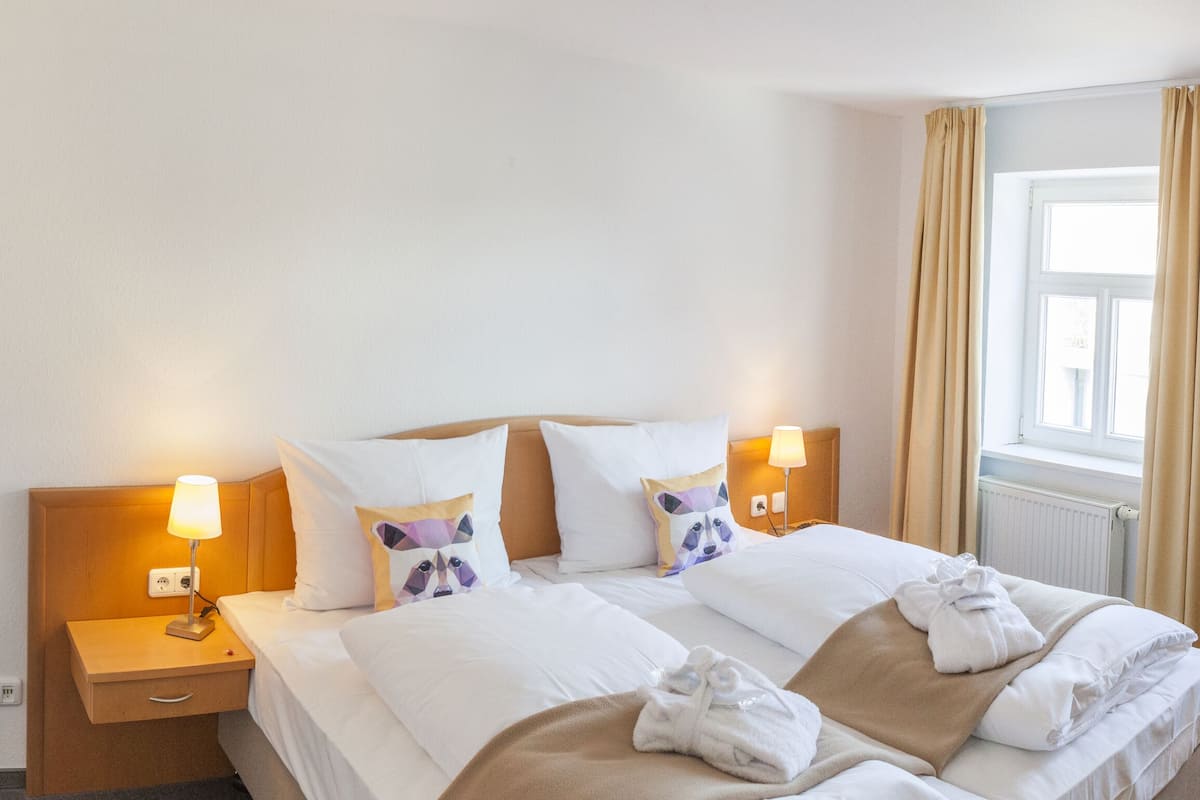 A hotel room with a bed, bedside lamps, a nightstand, a window with curtains, and a wall with a painting.