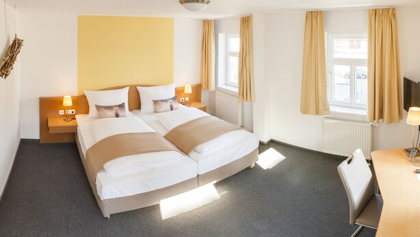 Habitación de hotel con cama doble, dos mesitas de noche con lámparas, un escritorio con silla, un ventanal con cortinas y un televisor montado en la pared.