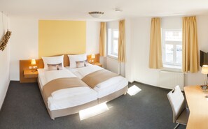 Habitación de hotel con cama doble, dos mesitas de noche con lámparas, un escritorio con silla, un ventanal con cortinas y un televisor montado en la pared.