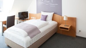 A hotel room with a bed, a desk with a TV, a lamp, and a wall with a decorative sign. - Hotel am Markt (Hungen)