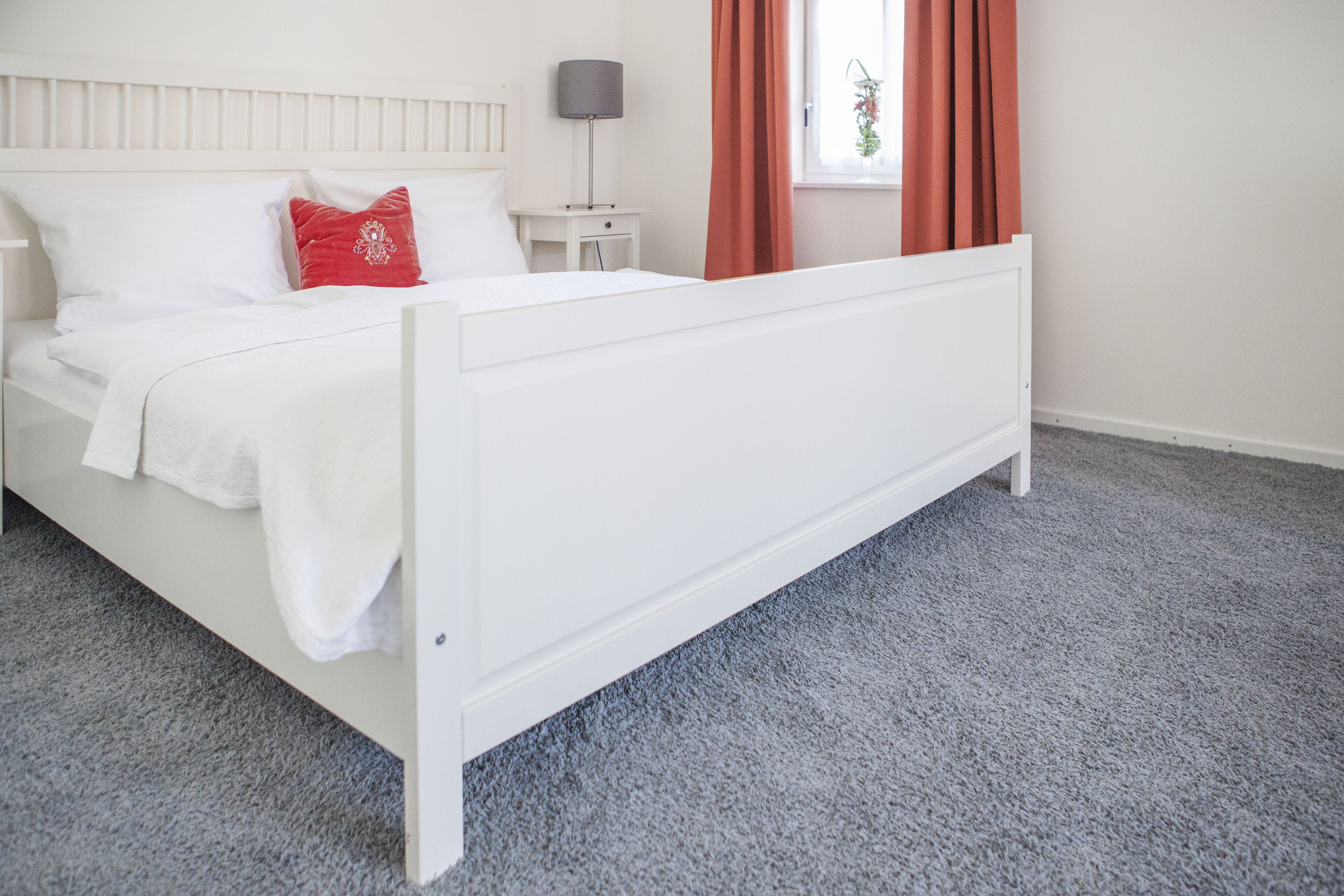 Una cama blanca con un almohadón rojo, una mesita de noche con una lámpara y una ventana con cortinas.