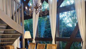 A wooden dining table with six chairs, a chandelier, and a view of greenery through large windows. - Landhaus Szczecin Lagoon, house3 (Dargen)