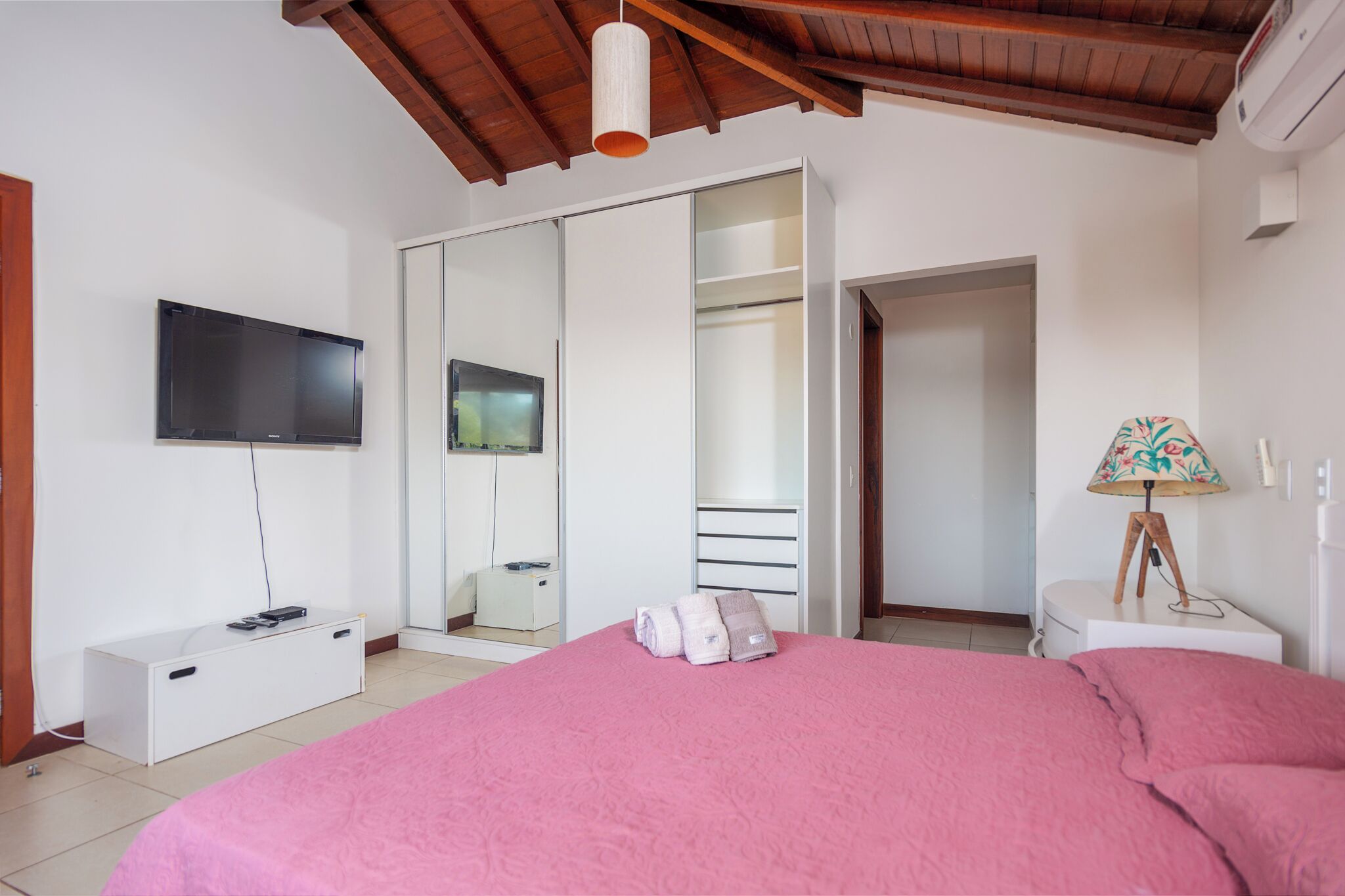 Een slaapkamer met een groot bed, een aan de muur bevestigde televisie, een kledingkast en een nachtkastje met een lamp.