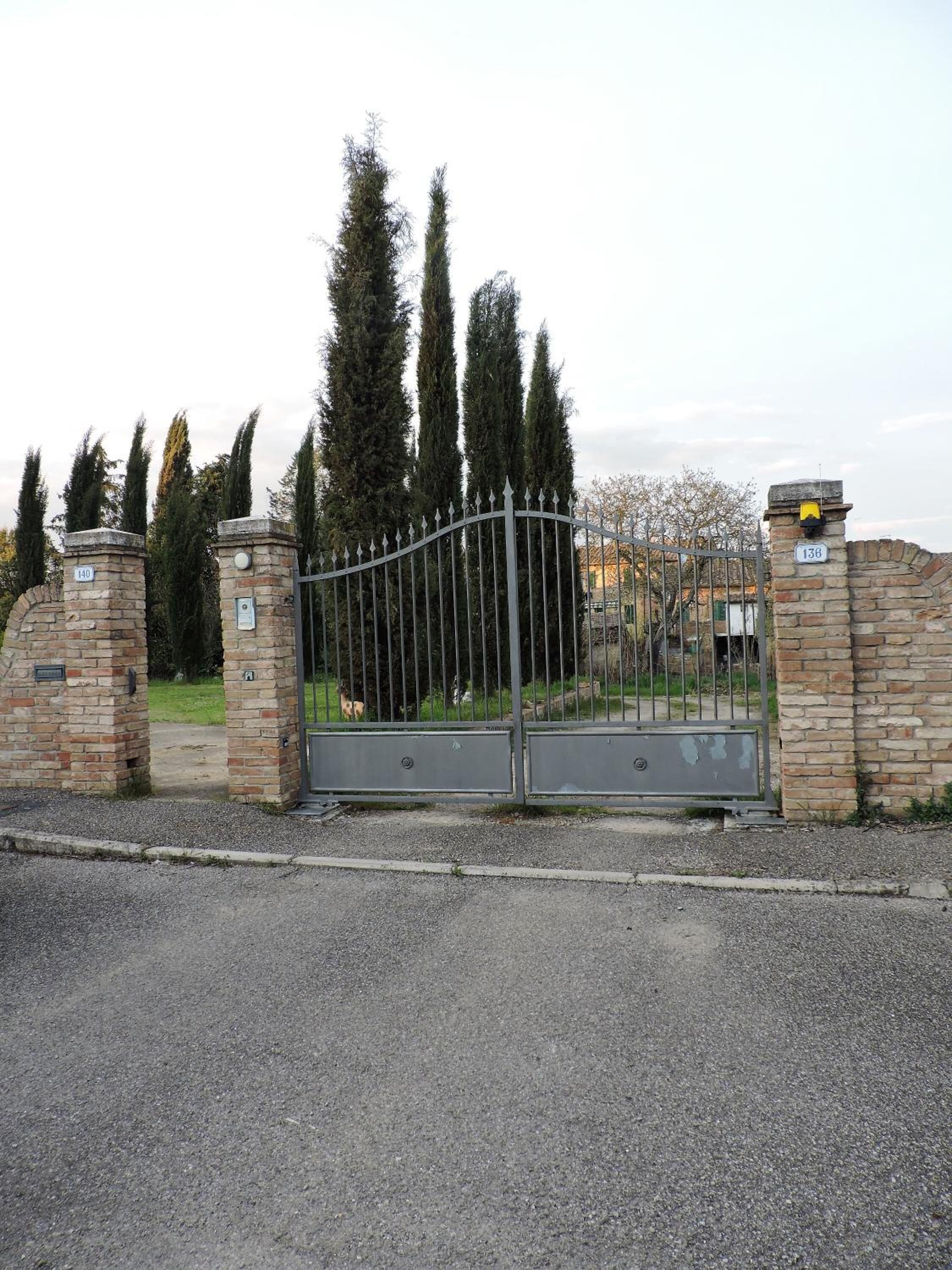 A gated entrance with stone pillars and a metal gate, leading to a building with a brick wall and a number sign.