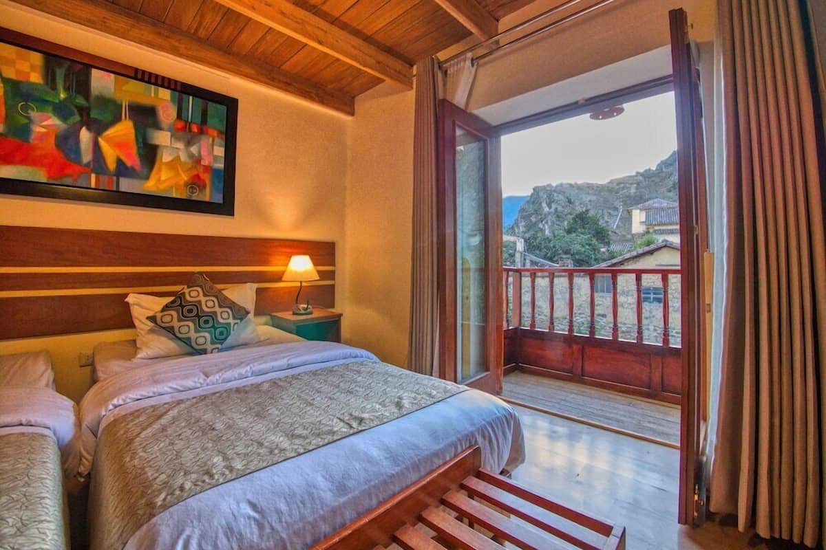 Un dormitorio con cama, mesita de noche, lámpara, un cuadro y balcón con vistas a edificios y una montaña.