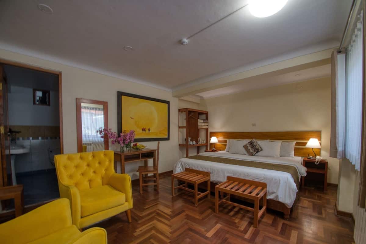 Habitación de hotel con cama, dos mesitas de noche, un escritorio con silla, un sillón amarillo y un cuadro grande en la pared.