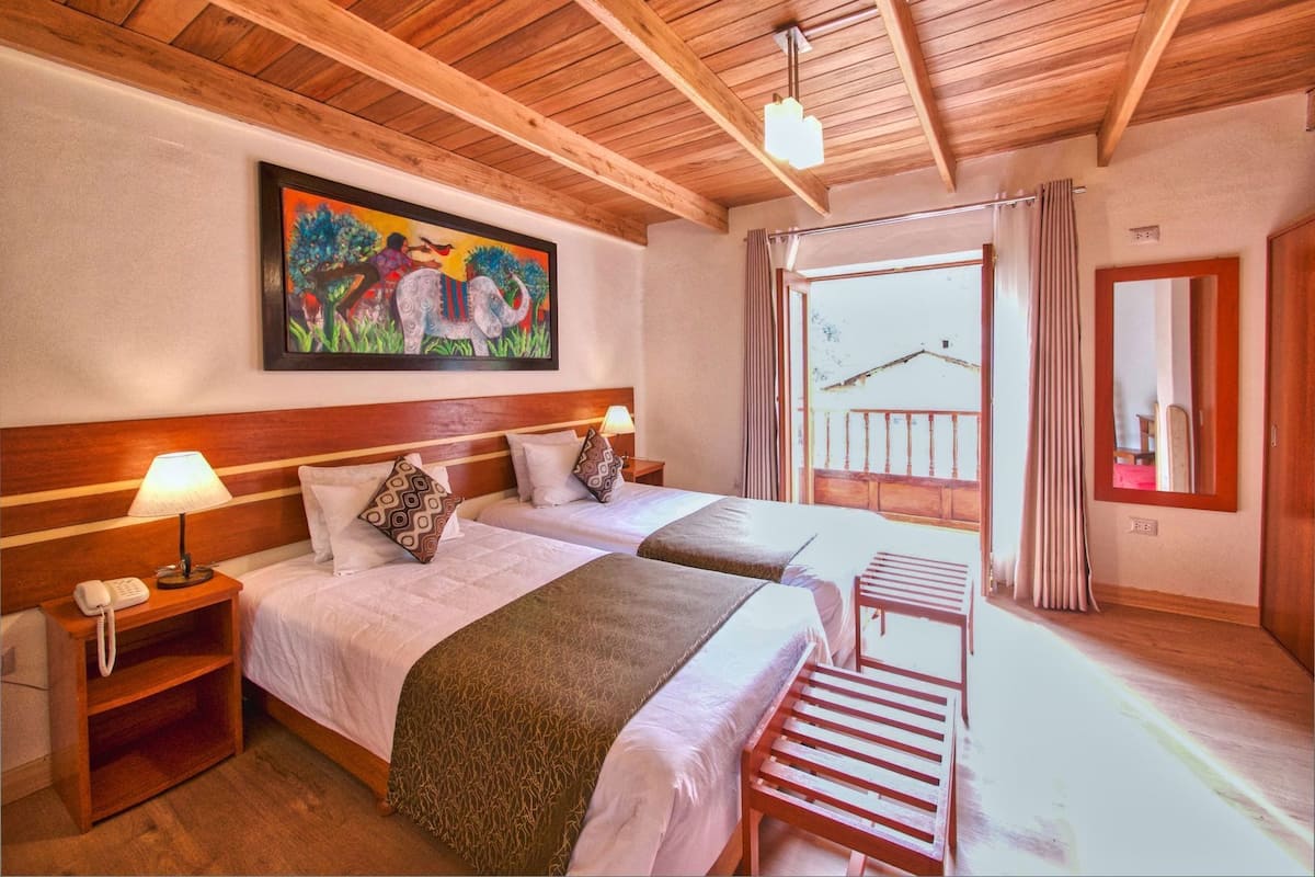 Habitación de hotel con dos camas, cabeceros de madera, un cuadro en la pared, un balcón con cortinas y una silla de madera.