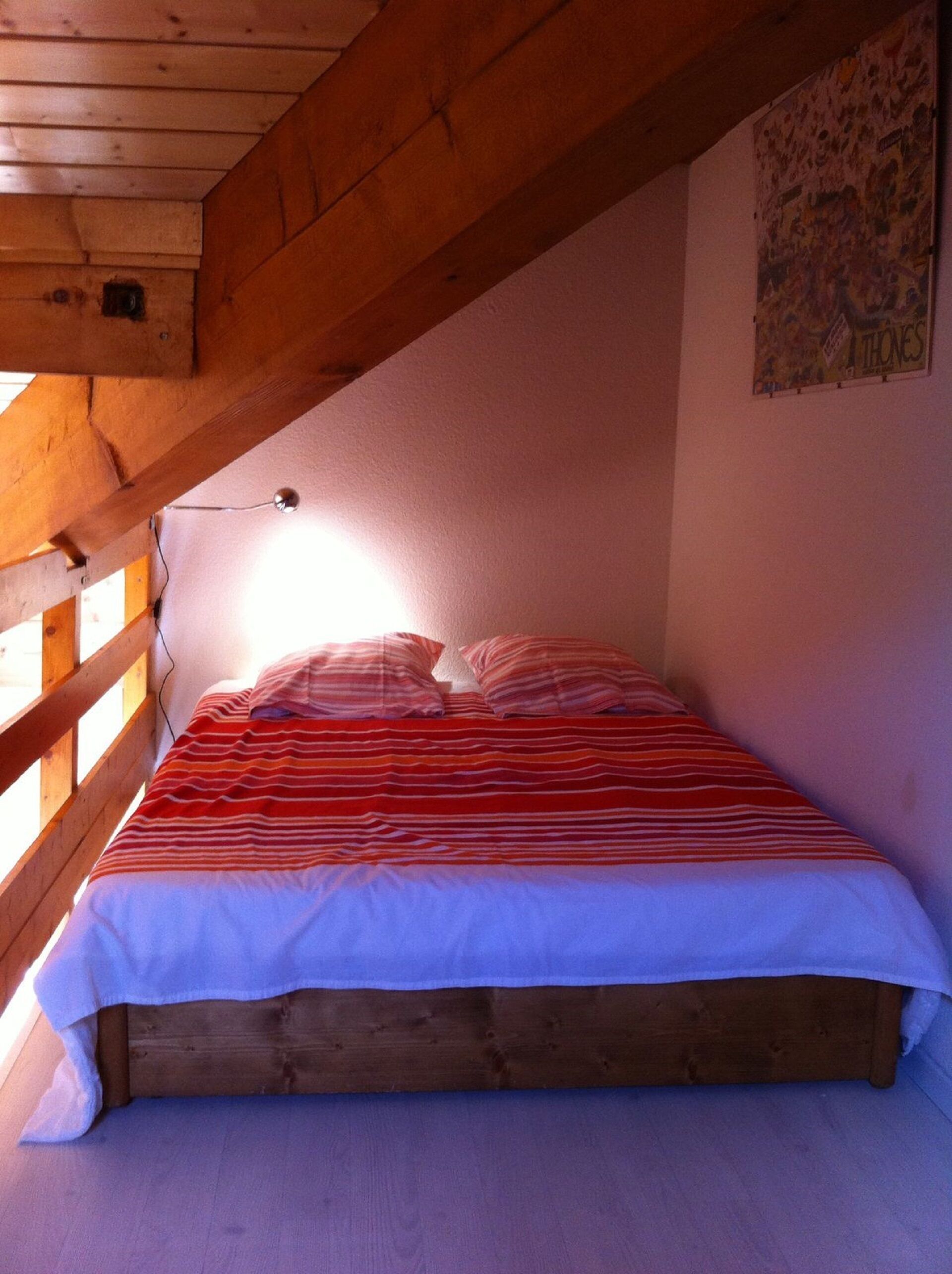 A single bed in a room with wooden beams and a sloped ceiling, a wall-mounted light, and a poster.