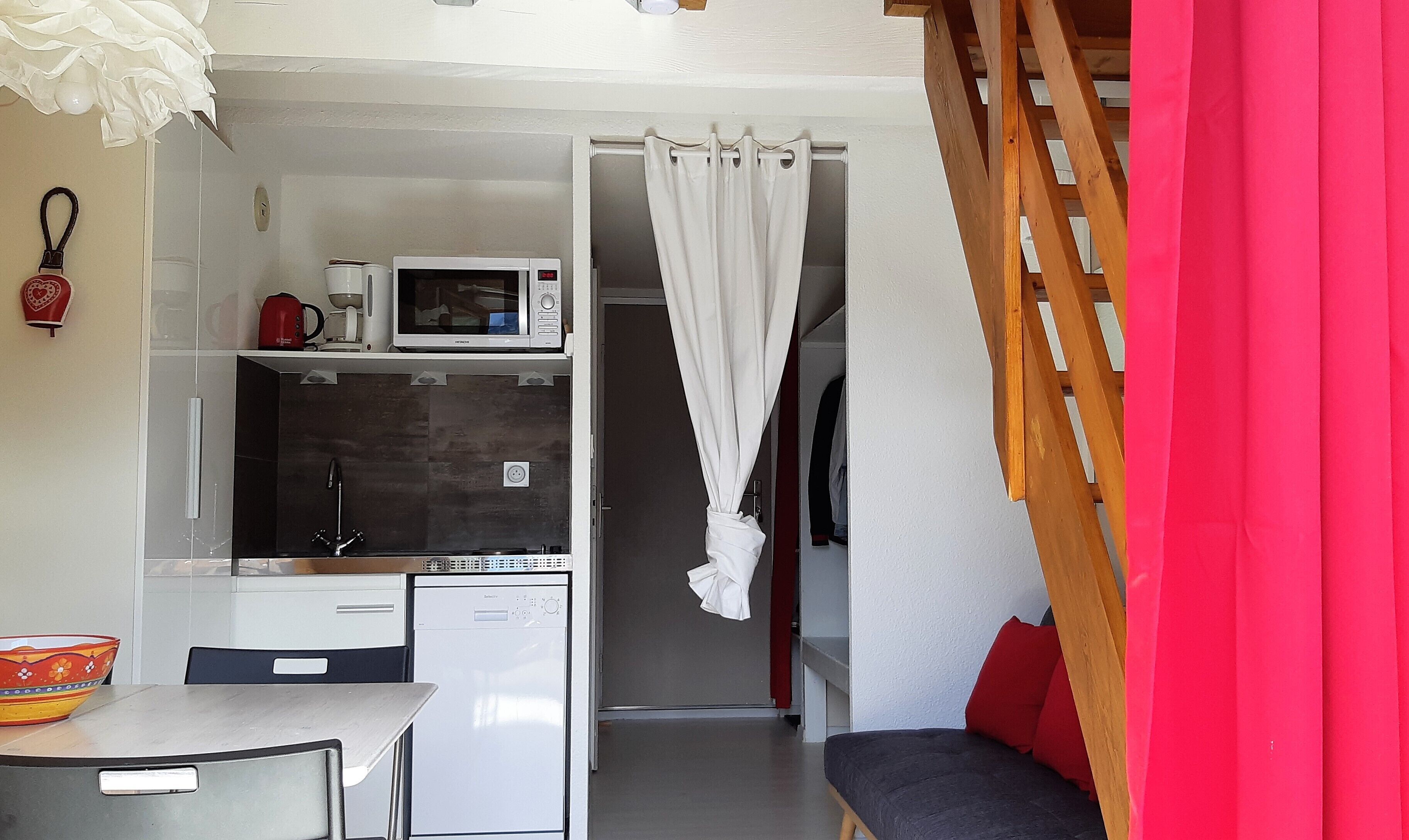 A compact kitchen with a microwave, coffee maker, and dishwasher. There is a small dining area with a table and chairs. A red curtain and a wooden ladder are visible.