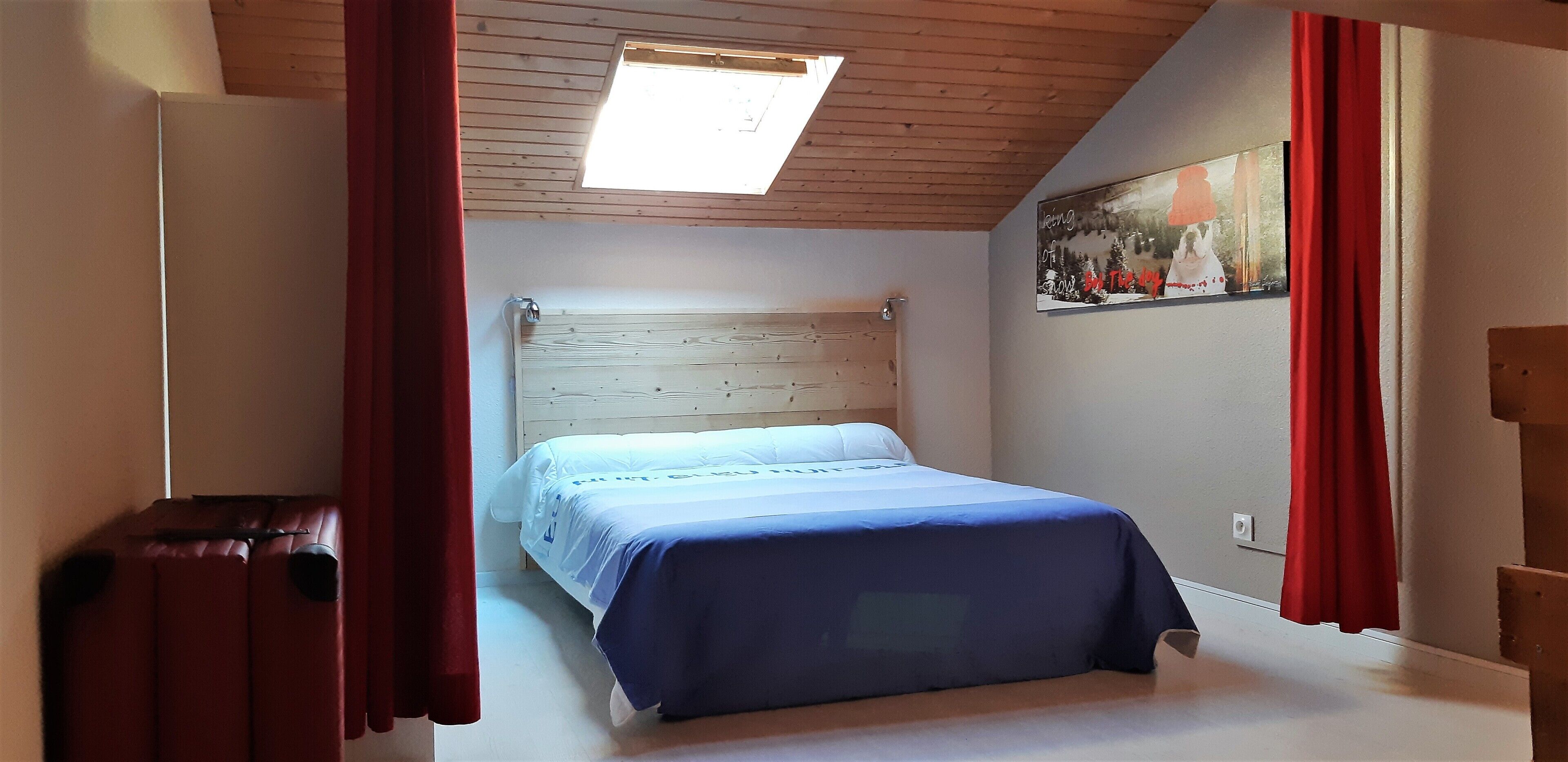 Un dormitorio con techo de madera, una cama con cubrecama azul, una cortina roja y un cuadro en la pared.