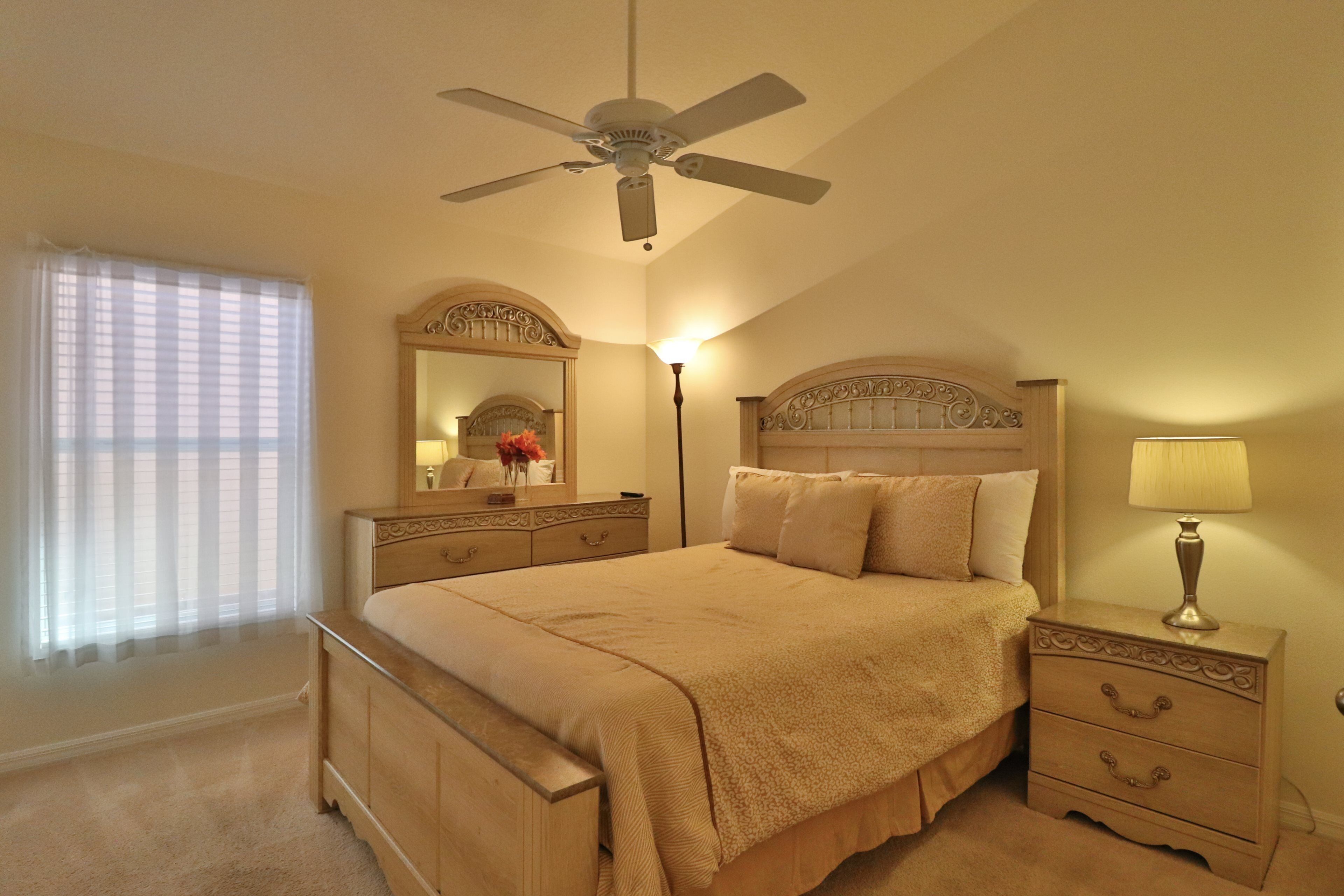 A bedroom with a bed, nightstand, dresser, ceiling fan, and a window with sheer curtains.