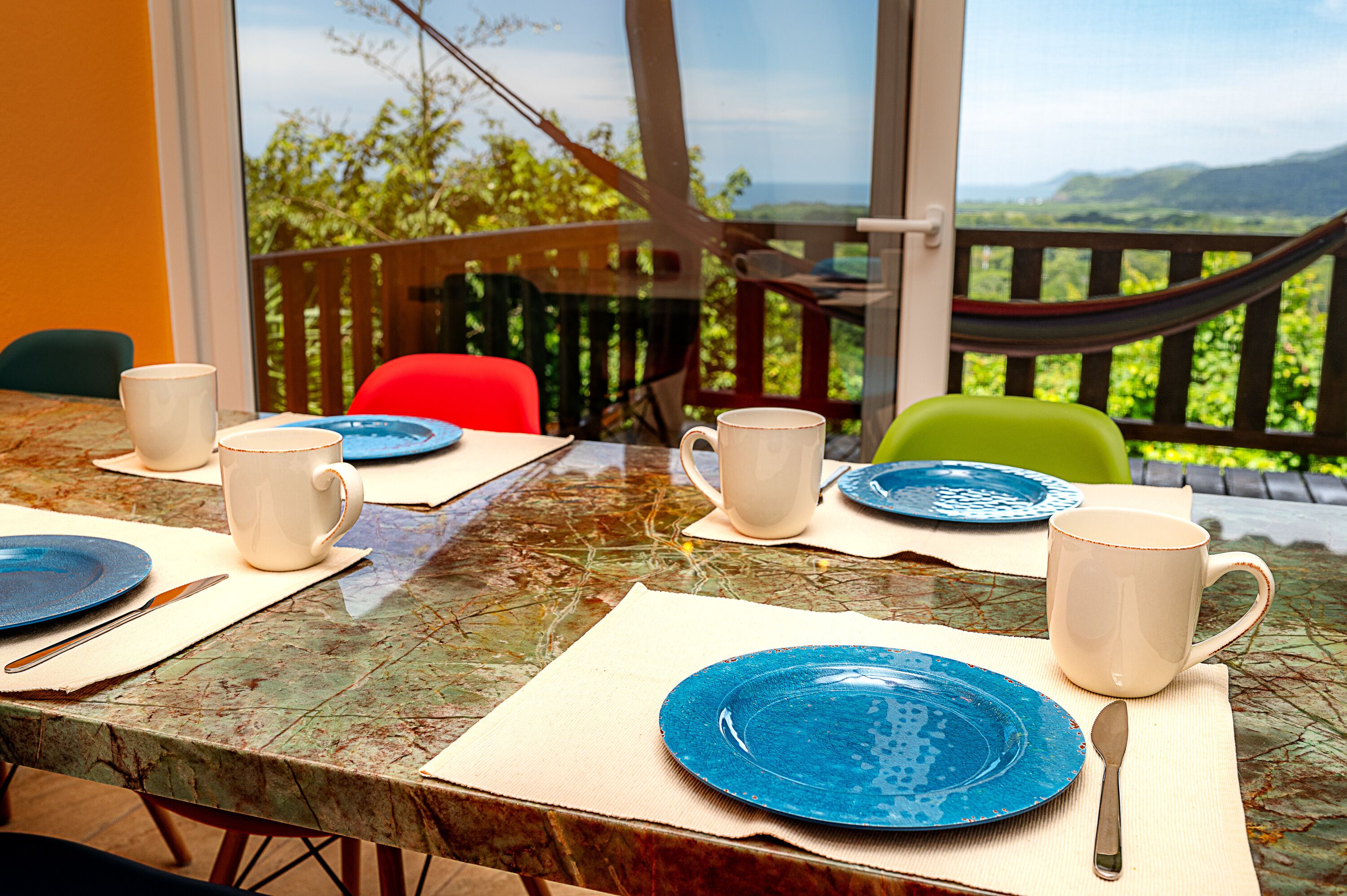 A dining table set with plates, cups, and cutlery, with a view of greenery and hills through a window.
