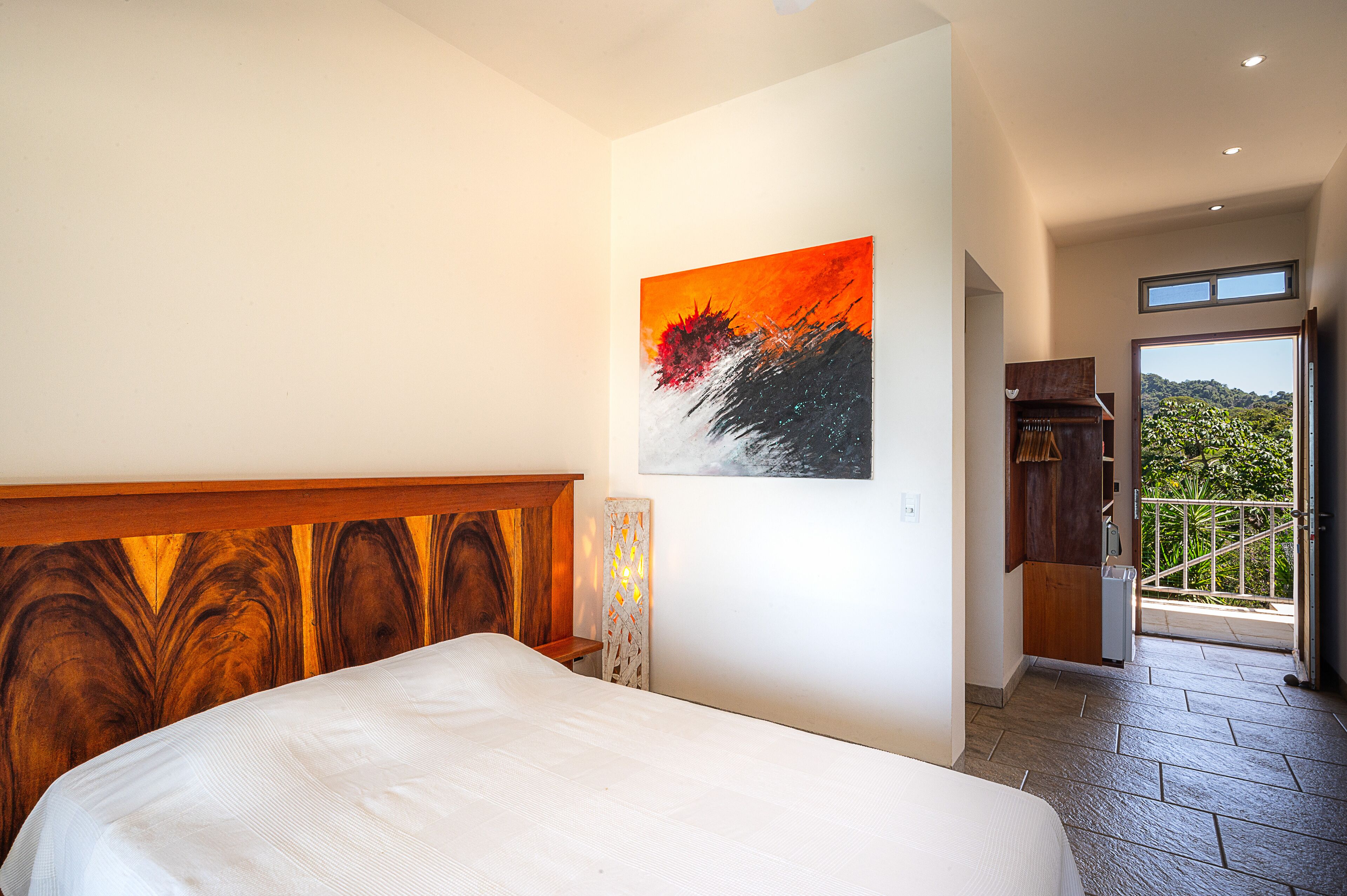 A bedroom with a wooden headboard, a painting on the wall, and a view of the outdoors through a door.