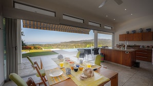 A dining area with a table set for breakfast, a kitchen with wooden cabinets, and a view of the landscape through large windows. - Casa Laureles (Bejuco)
