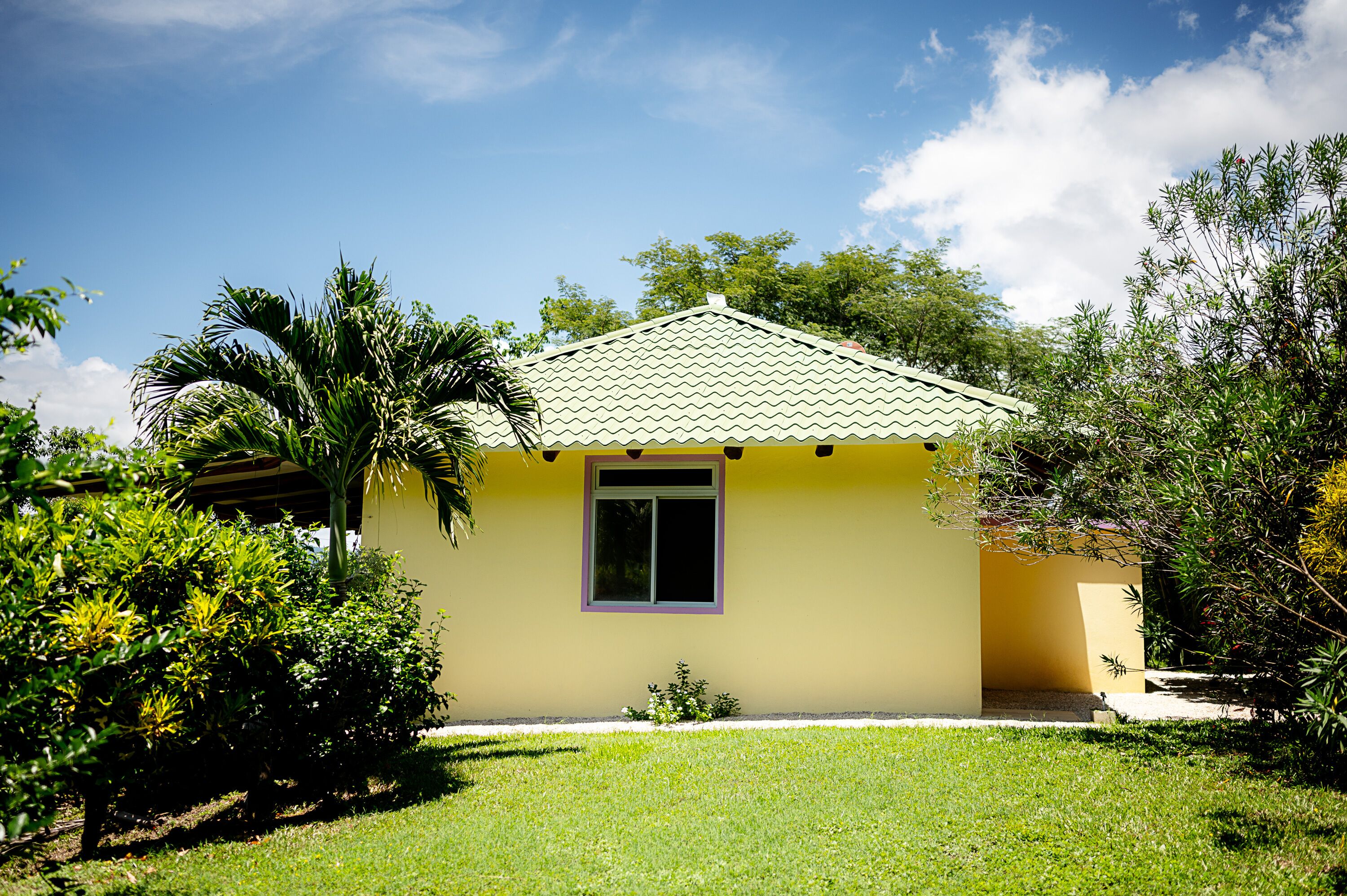 A small yellow house with a tiled roof, surrounded by lush greenery and a clear blue sky.