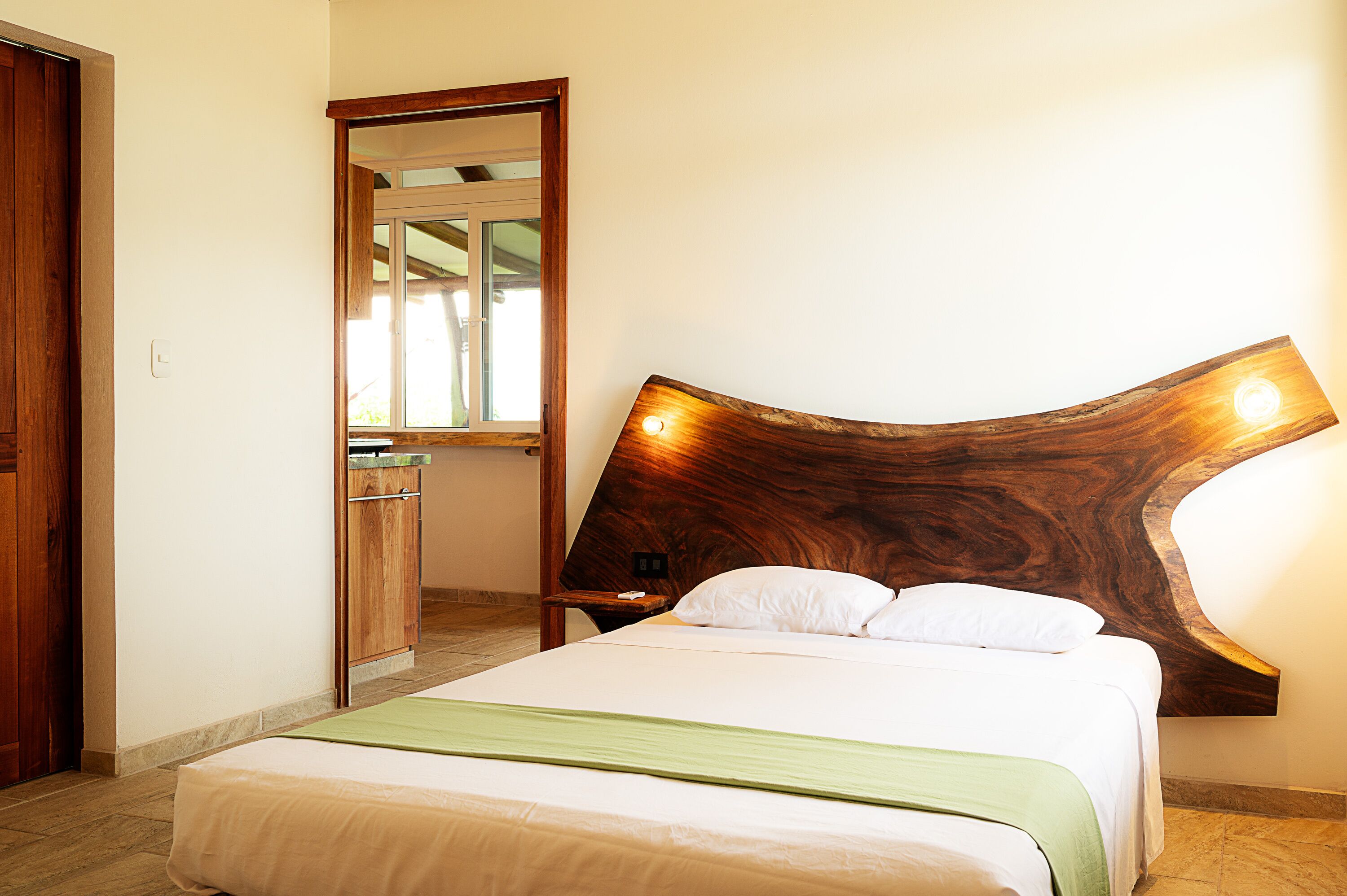 Un dormitorio con cabecera de madera, cama con ropa de cama blanca, manta verde y mesita de noche de madera.