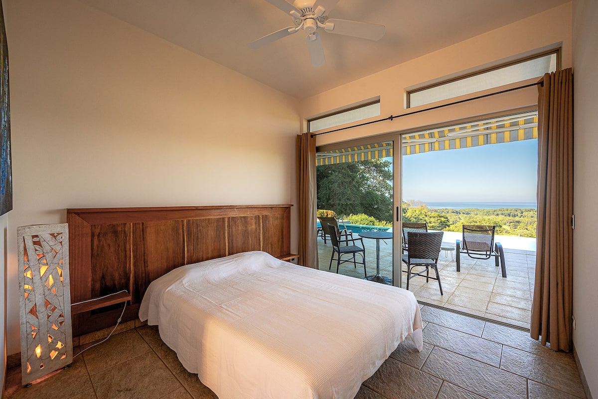 Ein Schlafzimmer mit einem großen Bett, einem Deckenventilator und Blick auf eine Terrasse mit Gartenmöbeln.