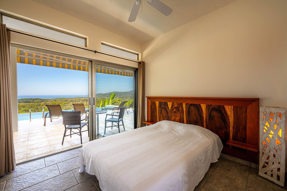 Ein Hotelzimmer mit Bett, Meerblick, einer Terrasse mit Stühlen und einem Deckenventilator.