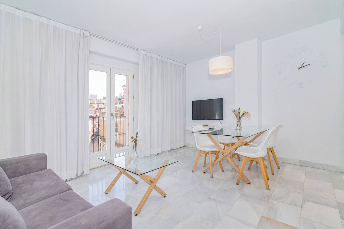 Una sala de estar moderna con una mesa de centro de vidrio, un sofá gris, un comedor con sillas blancas, un televisor y un amplio ventanal con cortinas.