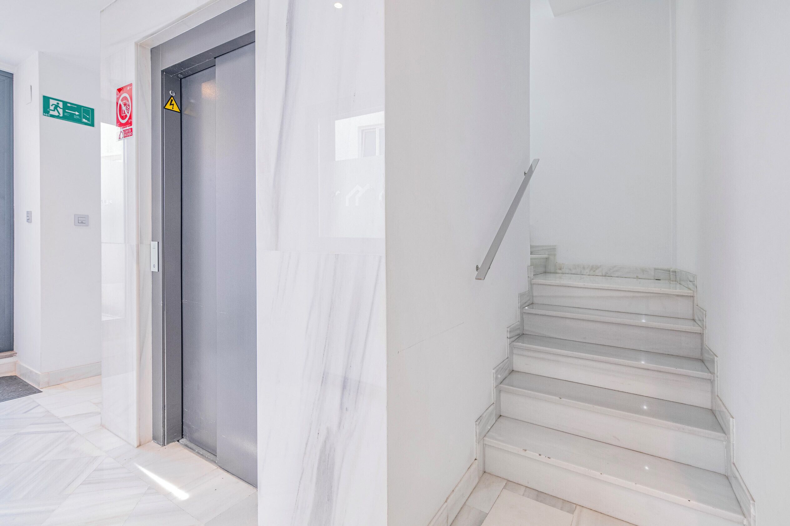 Un interior moderno con una escalera de mármol, un ascensor cerrado y señalización direccional.