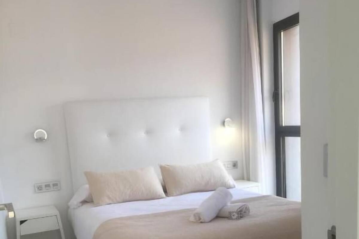 Un dormitorio ordenado con cama, mesita de noche y ventana con cortinas.
