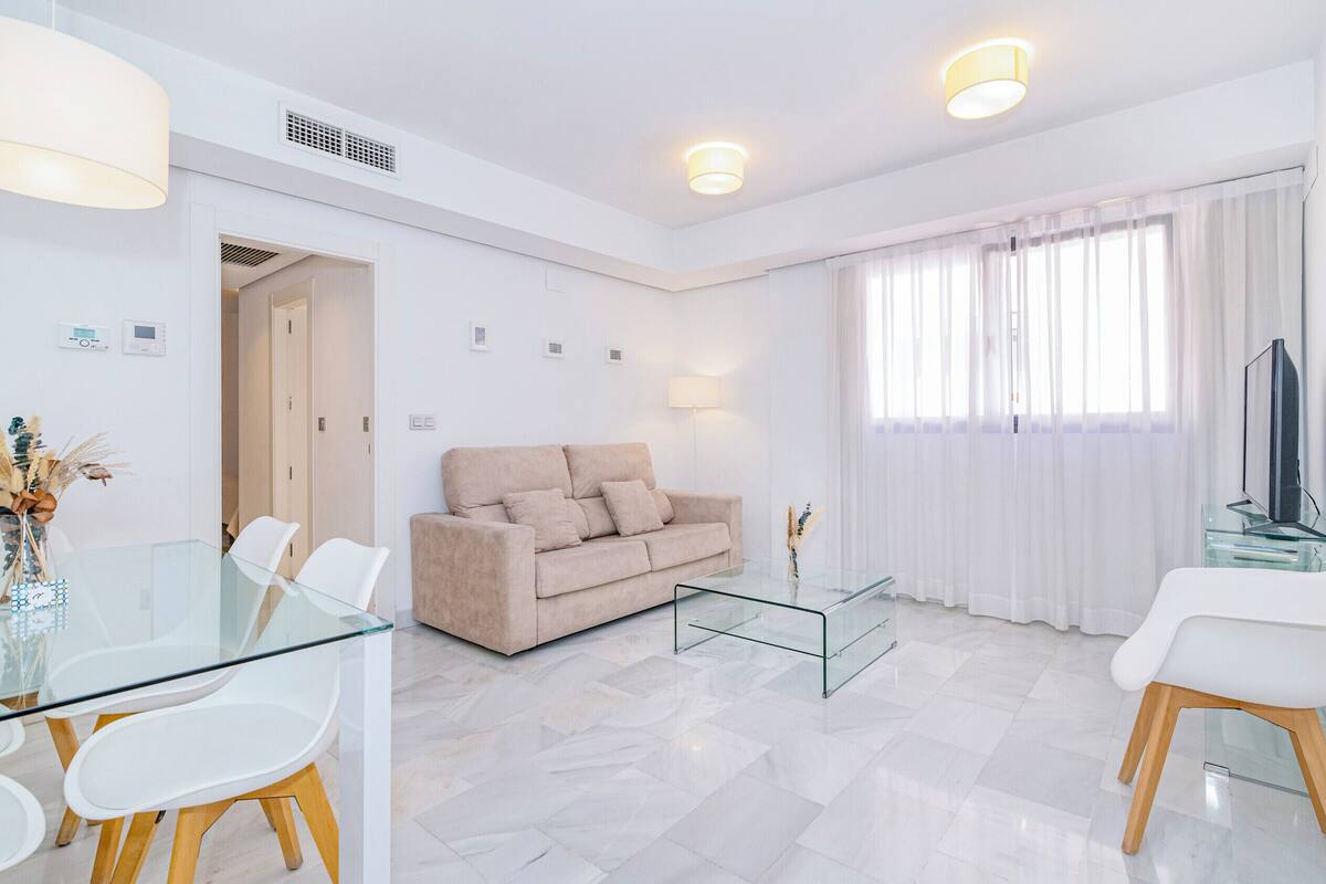 Una sala moderna con una mesa de centro de vidrio, sofá color beige, sillas blancas y un televisor.