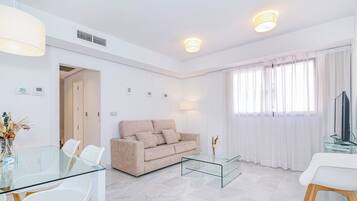 Una sala moderna con una mesa de centro de vidrio, sofá color beige, sillas blancas y un televisor.