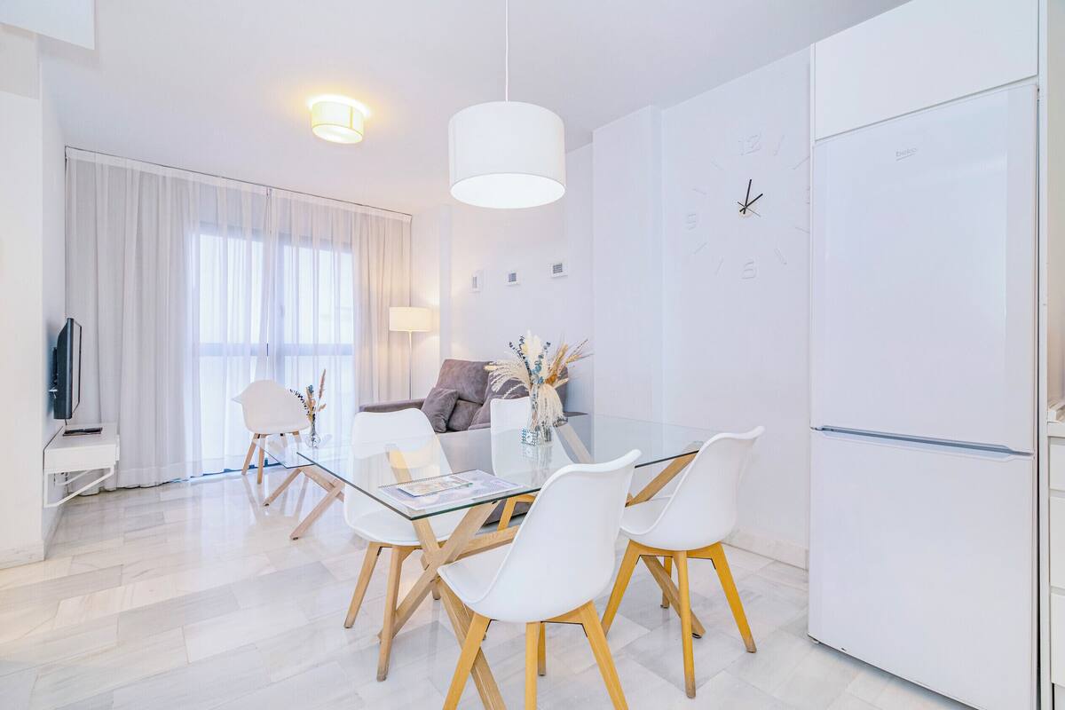 Un comedor moderno con una mesa de vidrio, sillas blancas, un refrigerador blanco y un televisor al fondo.