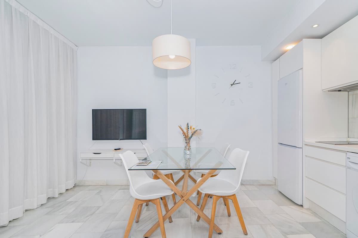 Una cocina moderna con armarios blancos, horno empotrado y comedor con mesa de vidrio y sillas blancas.