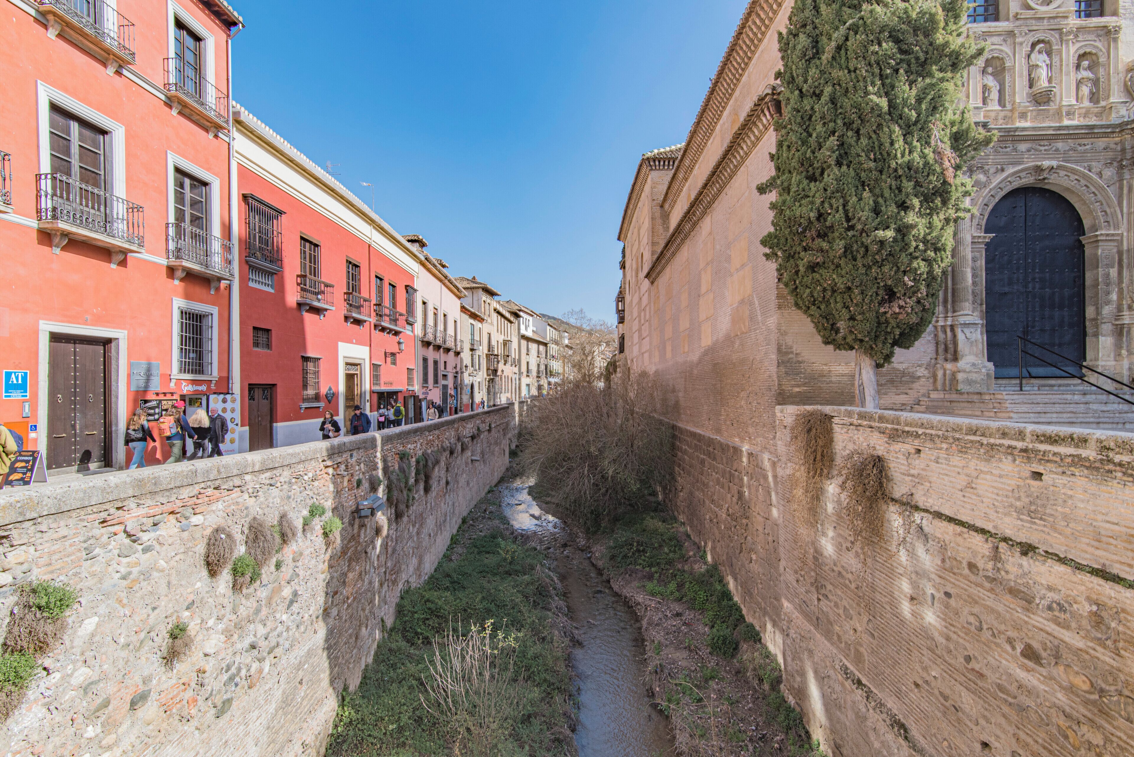 A narrow urban street with buildings on either side, a small water channel, and a stone wall.