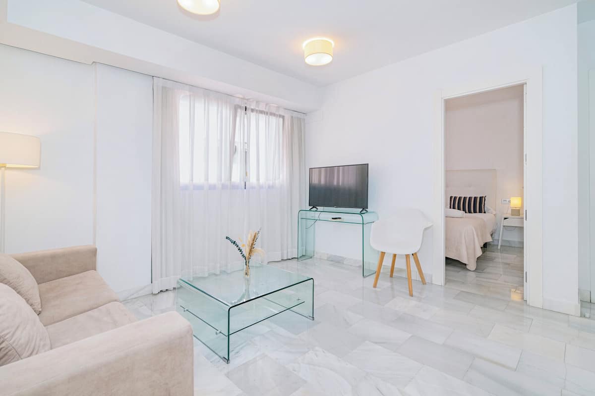 Una habitación de hotel moderna con una mesa de centro de vidrio, un sofá, un televisor sobre un escritorio de cristal y una cama con edredón a rayas.