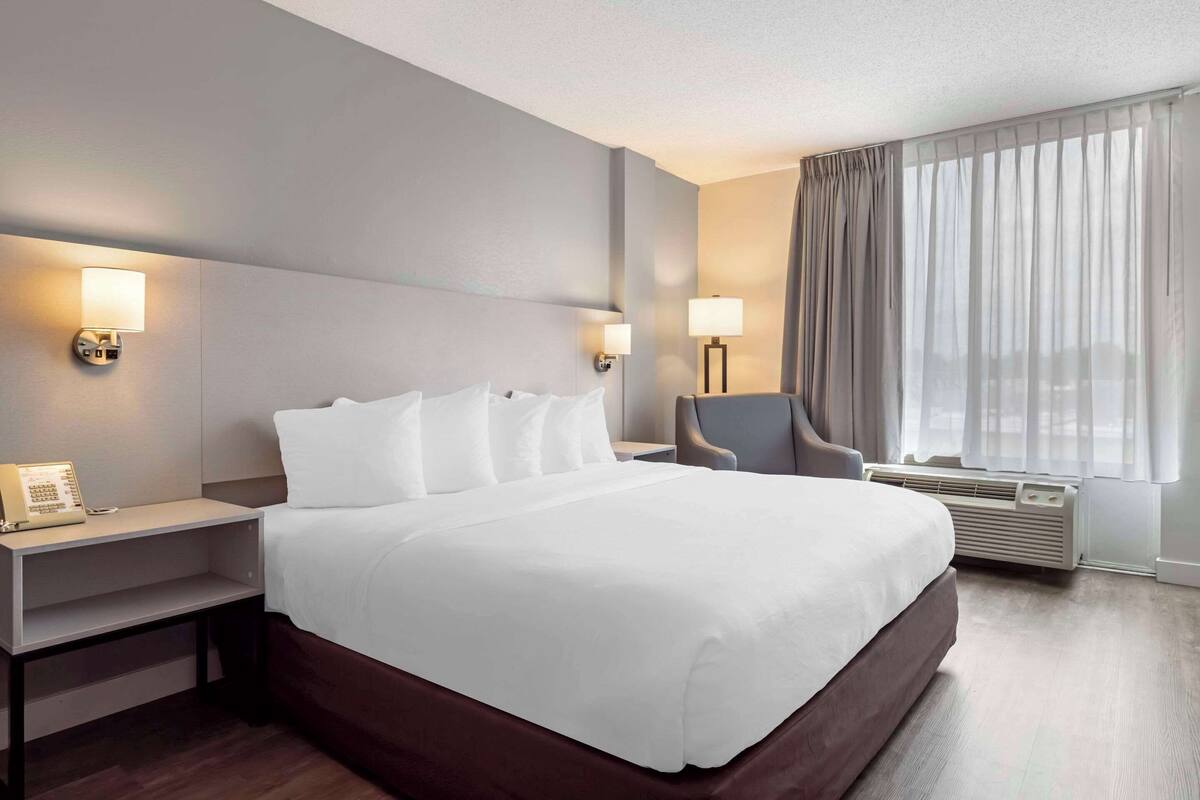 A hotel room with a large bed, bedside lamps, a chair, a desk, and a window with curtains.