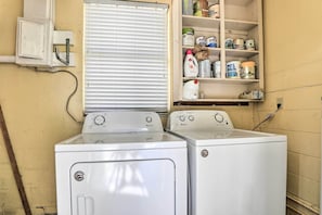 Un lavadero con dos lavadoras de carga frontal, un estante con diversos productos de limpieza y una ventana con persianas.