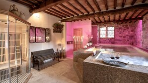 Une pièce aux murs de pierre, avec un banc, un espace clos en verre et un coin réception comprenant un jacuzzi et deux verres de vin rouge.