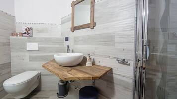 Un baño con tocador de madera, lavamanos blanco, espejo, inodoro y zona de ducha.