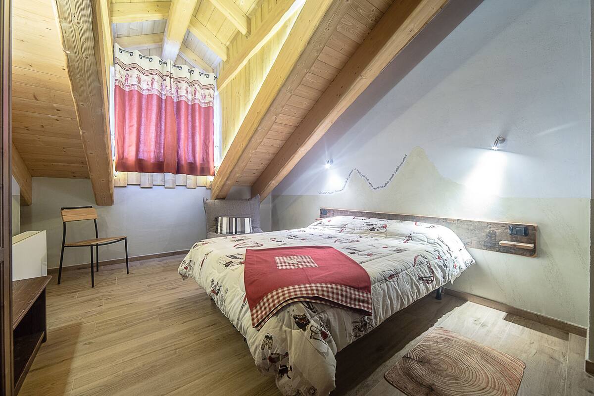 Une chambre avec un plafond en pente, des poutres en bois, un lit recouvert d’une couvre-lit à motifs, une chaise et une petite table.